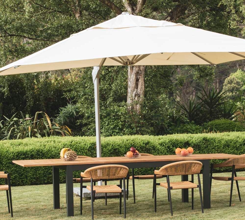 Outdoor dining area with large beige umbrella, wooden table, wicker chairs, fruits on table, surrounded by garden greenery