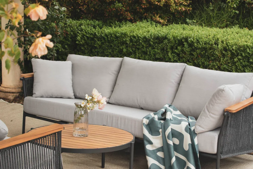 Outdoor patio furniture setup featuring a grey sofa, wooden - accented armrests, a round coffee table, a vase of flowers, and a draped throw blanket, surrounded by garden greenery.