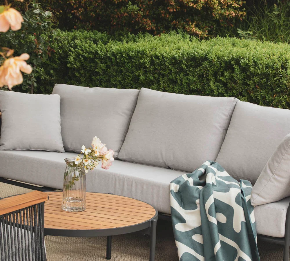 Outdoor patio furniture setup featuring a grey sofa, wooden - accented armrests, a round coffee table, a vase of flowers, and a draped throw blanket, surrounded by garden greenery.