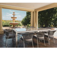Outdoor dining area with a large rectangular table, woven chairs, set under a covered patio, featuring a decorative fountain and lush greenery in the background.