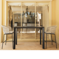 Outdoor bar table with two woven bar stools, set in front of large sliding glass doors showing an interior kitchen and chandelier.