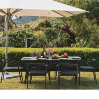 Outdoor dining set with a large beige umbrella, black table, black rope chairs, tableware, fruits, a bottle, and a flower vase, set in a lush garden with greenery in the background.
